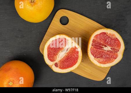 Gruppe von zwei ganze zwei Hälften von frischem rosa Grapefruit auf Bambus Schneidebrett flatlay am grauen Stein Stockfoto