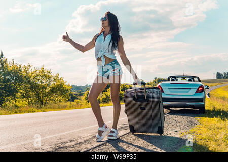 Frau mittleren Alters mit einer Reisetasche per Anhalter Fahren auf schmalen leere Landstraße, im Sommer auf dem Hintergrund einer kaputten Auto schöne Natur außerhalb der Stadt Stockfoto