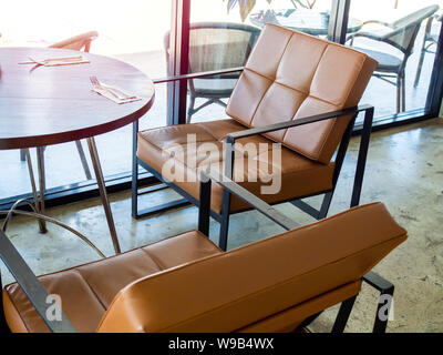 Black Metal frame und tan Leder Sessel Retro Stil und runden Holztisch in der Nähe von Glas Wand im Cafe. Stockfoto