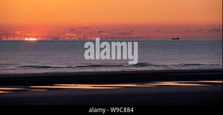 Ein ruhiger Sonnenuntergang über der Irischen See, von einem Strand mit Mustern im Sand und einem Windpark am Horizont mit der untergehenden Sonne. Stockfoto