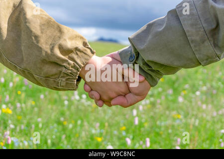 Die Reisenden der Mann und die Frau in der rauhen camping Outfits sind halten sich an den Händen (nur Hände sind in der Nähe gesehen) vor dem Hintergrund einer unscharfen wilde Wiese ein Stockfoto