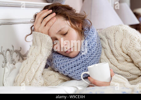 Traurig allein junge Frau im weißen Pullover und blauen Schal Gefühl, Kopfschmerzen, Kälte krank und Ruhe zuhause im Bett. Halten Sie ihren schmerzlichen Kopf und denken, was Stockfoto
