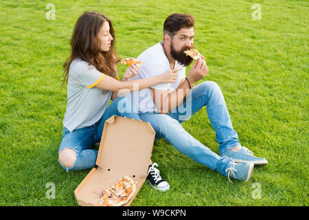 Paar essen Pizza entspannen auf grünen Rasen. Fast Food Delivery. Bärtiger Mann und Frau genießen cheesy Pizza. Paar in Liebe dating im Freien mit Pizza. Hungrige Studenten gemeinsame Essen. Genuss pur. Stockfoto