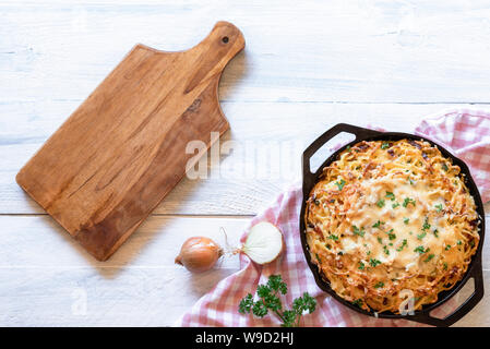 Käse Nudeln in Gusseisen Pfanne und leer Schneidbrett auf einem weißen Tisch. Oben Ansicht der Deutschen traditionellen Käse Spätzle mit Speck. Schwäbische Küche. Stockfoto