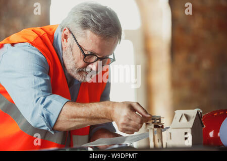 Close up photoshot der männlichen Architekt - Ingenieur ein Modell der Zukunft Haus für die junge Familie. Mann bei der Arbeit im Büro mit Miniatur, Zeichnungen, Blaupause. Erstes Haus, Industrie, Gebäude Konzept. Stockfoto