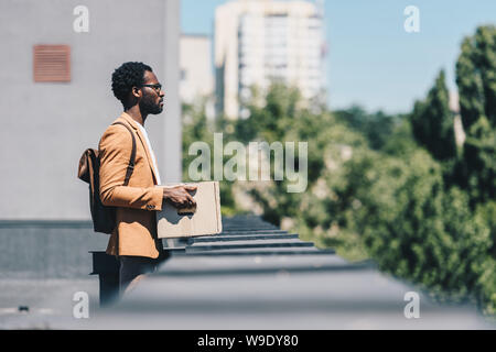 Selektiver Fokus der afrikanischen amerikanischen Geschäftsmann holding Karton und weg schauen beim Stehen auf dem Dach Stockfoto