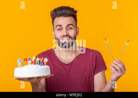Positive junge Mann hält ein Happy birthday cake und zwei brennende bengalische Lichter auf gelbem Hintergrund posiert. Stockfoto