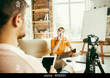 Zwei junge Kaukasier Männlich Blogger in Freizeitkleidung mit professioneller Ausstattung oder der Videokamera Video Interview zu Hause. Blogging, Videoblog, vlogging. Sprechen während Streaming live im Innenbereich. Stockfoto