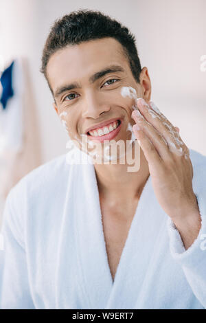 Hübscher junger Mann im Bademantel Anwendung Schaum im Gesicht und lächelt, während bei Camera suchen Stockfoto