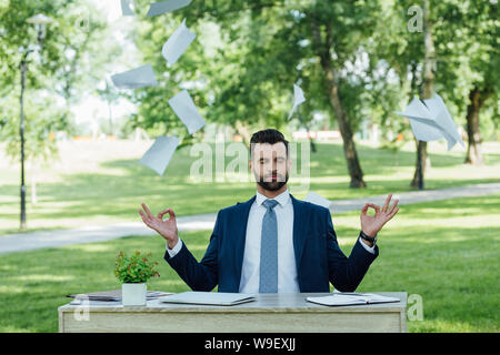 Junge Unternehmer im Park hinter der Tabelle und Meditieren in der Nähe von fliegende Papiere sitzen Stockfoto
