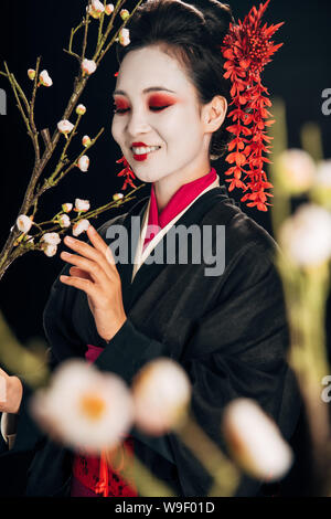 Selektiver Fokus der gerne schöne Geisha kimono in Schwarz mit roten Blumen im Haar und Sakura Niederlassungen isoliert auf Schwarz Stockfoto