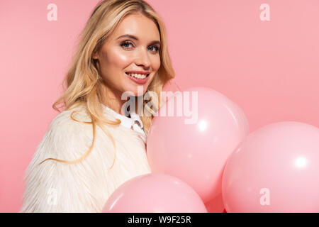 Lächelnd schöne blonde Frau in webpelzjacke Holding rosa Ballons isoliert auf Rosa Stockfoto