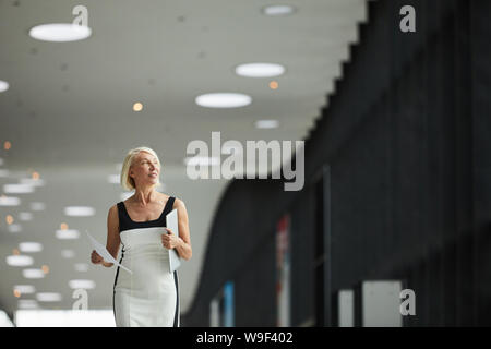 Reifen lächelnd Geschäftsfrau in elegantes Kleid mit Laptop und Dokumente und wandern durch das Büro Flur Stockfoto