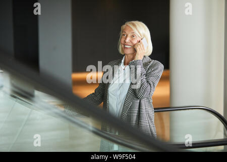 Reife Frau bewegte sich die Rolltreppe und Gespräch am Handy mit Lächeln Stockfoto