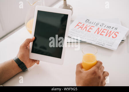 7/8-Ansicht der Mann hält ein Glas Orangensaft und digitale Tablet mit leerer Bildschirm beim Sitzen am Küchentisch in der Nähe von gefälschten Nachrichten Zeitung Stockfoto