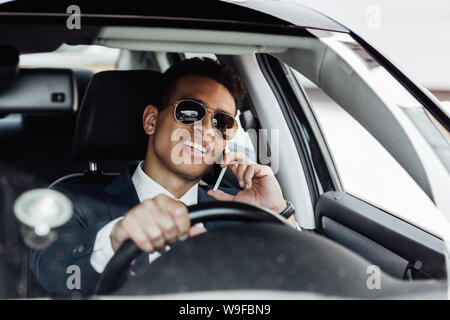 Afrikanische amerikanische Geschäftsmann in Anzug und Sonnenbrille fahren Auto und sprechen auf dem Smartphone an einem sonnigen Tag Stockfoto