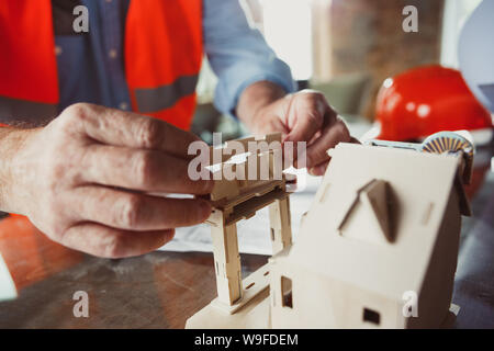 Close up photoshot der männlichen Architekt - Ingenieur ein Modell der Zukunft Haus für die junge Familie. Mann bei der Arbeit im Büro mit Miniatur, Zeichnungen, Blaupause. Erstes Haus, Industrie, Gebäude Konzept. Stockfoto