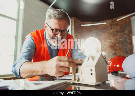 Close up photoshot der männlichen Architekt - Ingenieur ein Modell der Zukunft Haus für die junge Familie. Mann bei der Arbeit im Büro mit Miniatur, Zeichnungen, Blaupause. Erstes Haus, Industrie, Gebäude Konzept. Stockfoto