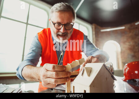 Close up photoshot der männlichen Architekt - Ingenieur ein Modell der Zukunft Haus für die junge Familie. Mann bei der Arbeit im Büro mit Miniatur, Zeichnungen, Blaupause. Erstes Haus, Industrie, Gebäude Konzept. Stockfoto