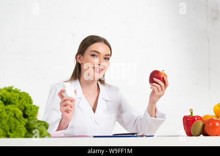 Lächelnd Ernährungsberater im weißen Mantel holding Pillen und Apple am Arbeitsplatz Stockfoto