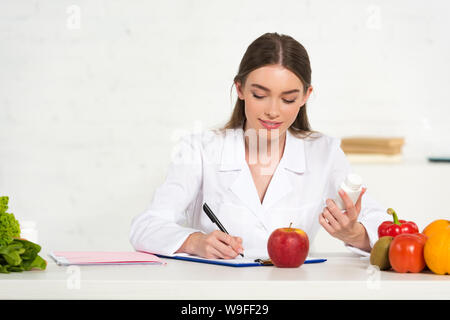 Lächelnd Ernährungsberater im weißen Mantel holding Pillen und Schreiben in die Zwischenablage am Arbeitsplatz mit Obst und Gemüse auf den Tisch Stockfoto