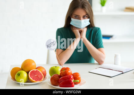 Traurige Frau in der medizinischen Maske propping Gesicht mit den Händen beim Sitzen am Tisch mit Obst, Gemüse und Schablone mit Inschrift Allergie Stockfoto