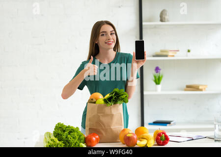 Lächelnde Mädchen in der Nähe von Papier Beutel mit Nahrung und Holding mit leeren Bildschirm zu Hause smartphone Stockfoto