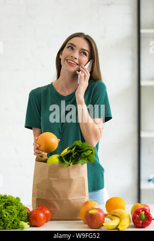 Lächelnde Mädchen in der Nähe von Papier Tüte mit frischen Lebensmitteln, orange und sprechen auf dem Smartphone zu Hause Stockfoto