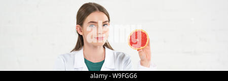 Panorama-aufnahme des Lächelns Ernährungsberater im weißen Mantel Holding schnitt Grapefruit und Kamera isoliert auf weißem Stockfoto