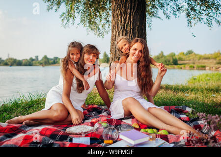 Familie mit Picknick im Sommer River bei Sonnenuntergang. Mutter, Großmutter und Kinder umarmen. Drei denerations Zeit miteinander zu verbringen Stockfoto