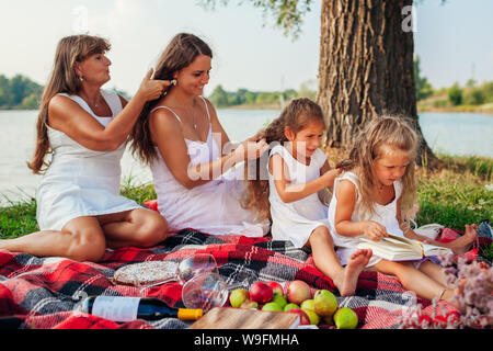 Mutter, Großmutter und Kinder weben Flechten, während die jüngsten Buch liest. Familie Spaß beim Picknick im Park. Drei denerations Stockfoto