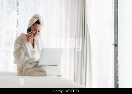 Entspannt Frau mit Bademantel mit Smartphone und Laptop im Bett Stockfoto