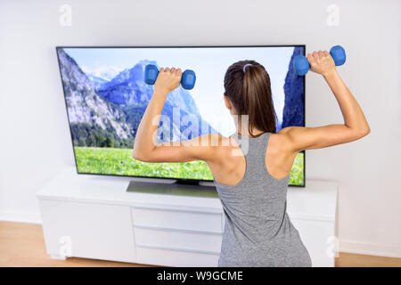 Fitness zu Hause - Frau vor dem Fernseher. Zurück einer jungen weiblichen Erwachsenen Fernsehen während Ihres Trainings, Gewichte zu Ton Arme und Schultern, nach einer Übung Video. Stockfoto