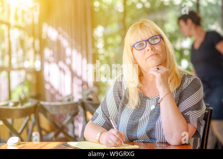 Porträt der Schönen plus größe Körper positive Frau arbeiten und sich Notizen zu ihrem Tagebuch an sonnigen Cafe. Elegante Dame mittleren Alters mit modernen Gläsern posieren. reife Frau Schriftsteller sitzen Schreiben. Stockfoto