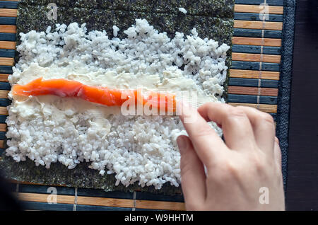 Die Sushi Rollen zu Hause: weibliche Hand Lachs Fisch Streifen auf Frischkäse, Reis, Nori Algen Blatt auf einem Bambus Matte gelegt. Ansicht von oben. Stockfoto