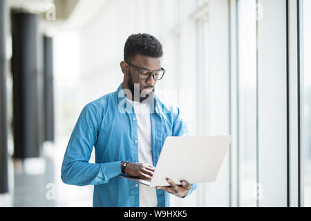 Liebe mein Job. Taille-up Portrait von positiven attraktive afrikanischer Mann in Gläsern steht in Büro und Betrieb moderner Laptop. Er ist auf der Suche nach Monitor Stockfoto