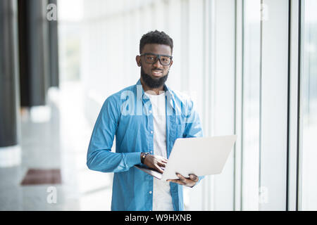 Liebe mein Job. Taille-up Portrait von positiven attraktive afrikanischer Mann in Gläsern steht in Büro und Betrieb moderner Laptop. Er ist auf der Suche nach Monitor Stockfoto