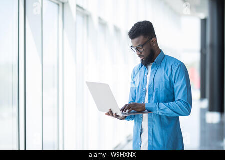 Liebe mein Job. Taille-up Portrait von positiven attraktive afrikanischer Mann in Gläsern steht in Büro und Betrieb moderner Laptop. Er ist auf der Suche nach Monitor Stockfoto