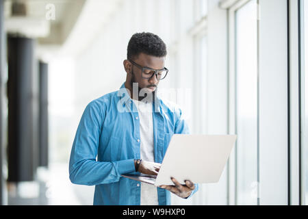 Liebe mein Job. Taille-up Portrait von positiven attraktive afrikanischer Mann in Gläsern steht in Büro und Betrieb moderner Laptop. Er ist auf der Suche nach Monitor Stockfoto