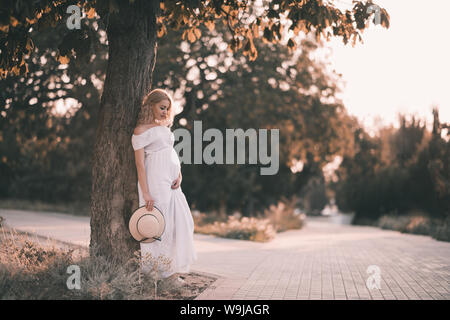 Schöne schwangere Frau 20-24 Jahre alten Standing in Park über Sonnenlicht. Das Tragen von eleganten weißen Kleid und mit Strohhut im Freien. Die Mutterschaft. Mater Stockfoto