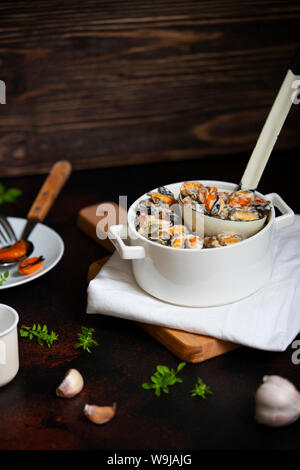 Muscheln in cremefarbenen und weißen Wein Sauce. Mit Knoblauch und Kräutern in einer weißen Wanne mit einer Pfanne serviert. Seafood gourmet Teller. Dunklen Hintergrund Stockfoto