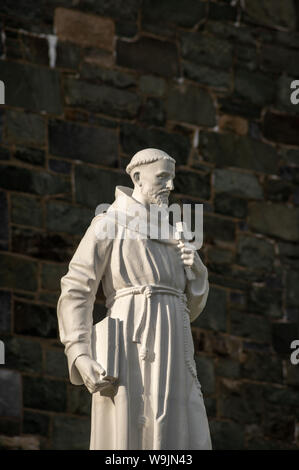 Religiöse Figur eines Heiligen Kreuz und Buch gegen Kirche stein Wand Fassade dunklen Hintergrund. Hl. Franz von Assisi Statue in Killarney, Irland. Stockfoto
