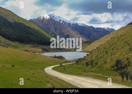 Der Blick hinunter eine unbefestigte Straße zu einem kleinen See, umgeben von Hügeln und Bergen, Schnee auf den Bergen, niemand im Bild Stockfoto