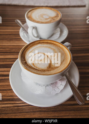 Zwei Tasse Cappuccino und Latte Kunst auf Holz- Hintergrund. Heißer Kaffee auf Tisch. Von der Seite. Schönen Schaum Stockfoto