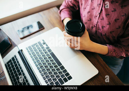 Überblick über eine junge Frau in einem Cafe vor dem Laptop arbeiten, mit einer Tasse Kaffee. Smartphone Neben. Stockfoto