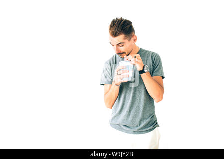 Glückliche junge verspielte Kerl in grauen T-Shirt mit einer Tasse Kaffee auf weißem Hintergrund isoliert, peeking im Inneren der Schale zu lächeln. Stockfoto