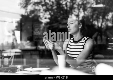 Nachdenklich kaukasische Frau mit Mobiltelefon, während Sie durch den Coffee shop Fenster während der Kaffeepause. Stockfoto