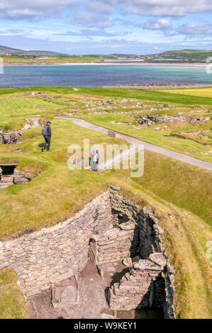 Touristen, die ein Steuerhaus bei der ausgegrabenen Stätte von jarlshof in der Nähe von Sumburgh Head im Süden von Mainland, Shetland. West Voe von sumburgh im Hintergrund. Stockfoto