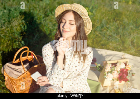 Junge hübsche, dunkelhaarige Mädchen in einem Strohhut bei Sonnenuntergang Picknick im Feld. Herbst. Camping Früchte und Getränke in einem Korb und Bouquet Stockfoto