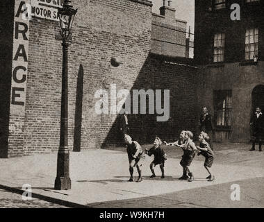 Circa 1940 - Britische Schule Kinder aus dem Norden Englands Fußball spielen in einem gepflasterten Platz neben der Straße, während die erwachsenen Nachbarn oder Väter auf. Stockfoto